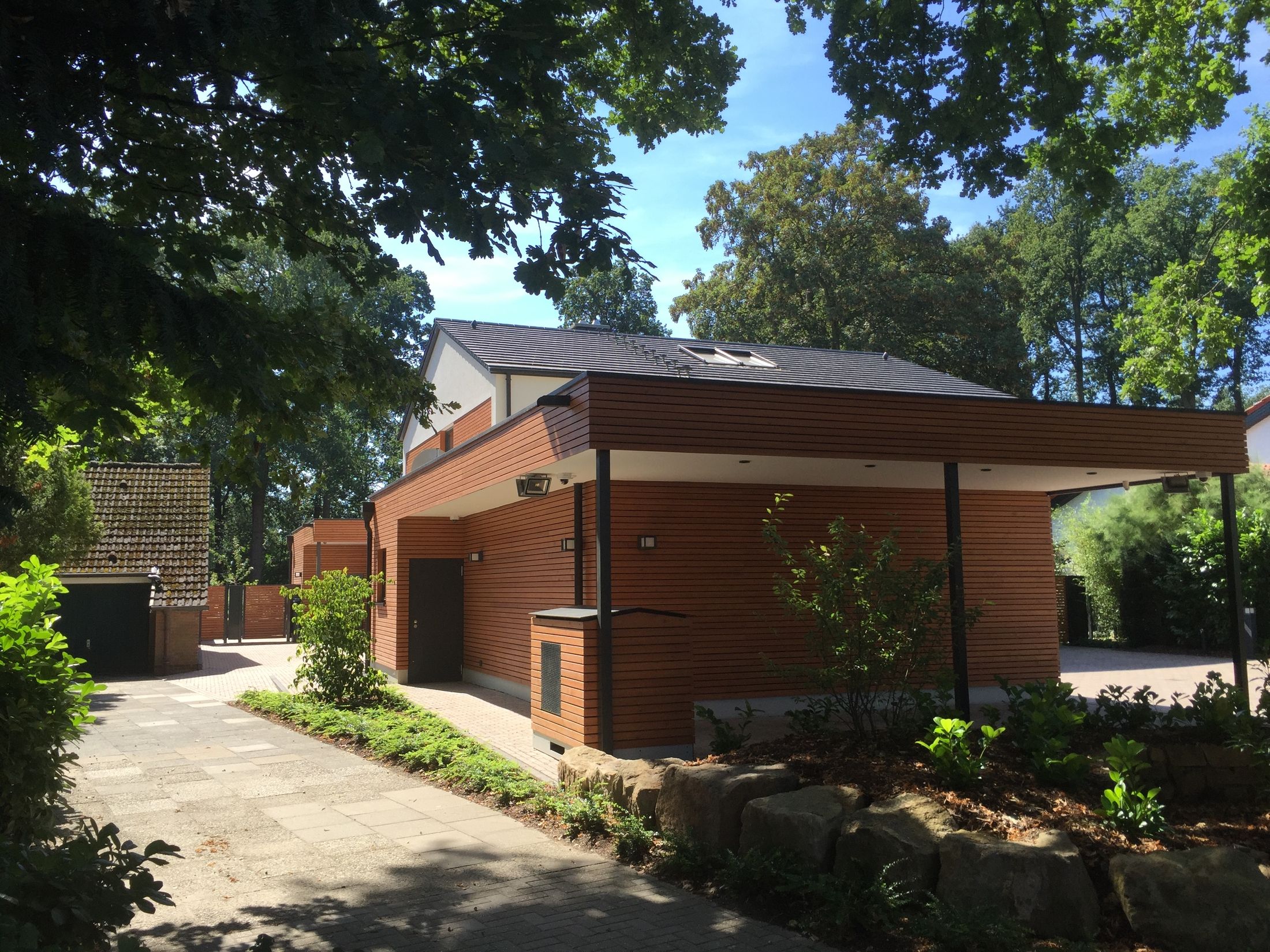 Modernes Haus mit Holzverkleidung, umgeben von Bäumen und einem gepflasterten Weg.